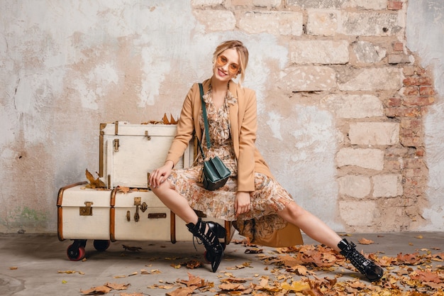 Attraente donna bionda alla moda in cappotto beige che si siede sulle valigie contro il muro in strada