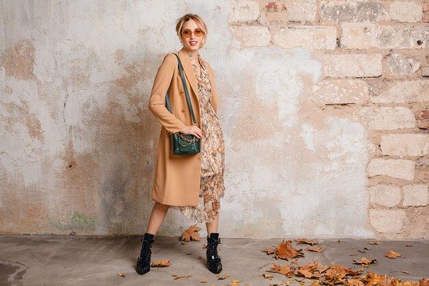 Attractive stylish blonde woman in beige coat walking in street against vintage wall