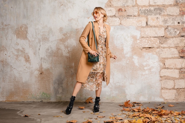 Attraente donna bionda alla moda in cappotto beige che cammina in strada contro il muro d'epoca