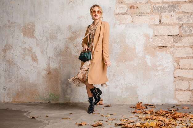 Attractive stylish blonde woman in beige coat walking in street against vintage wall