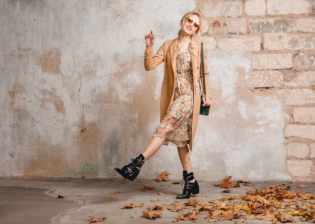 Привлекательная стильная блондинка в бежевом пальто гуляет по улице против старинной стены