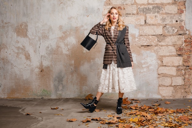 Attractive stylish blonde woman in checkered jacket walking against wall in street