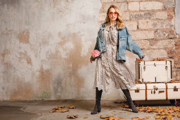 Attractive stylish blonde woman in jeans and oversize jacket walking against wall in street