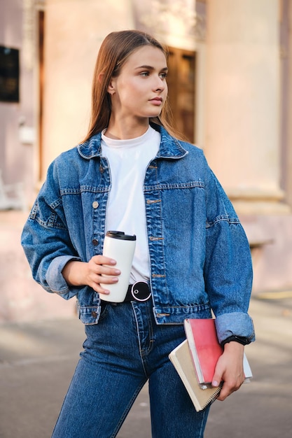 Attractive stylish casual student girl in denim jacket with coffee and textbooks thoughtfully looking away outdoor