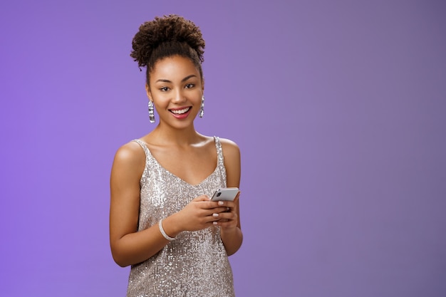 Привлекательная стильная уверенно афро-американская женщина в серебряном блестящем платье, держащая смартфон, обменивающийся сообщениями, прокручивая интернет-гаджет, глядя на счастливую счастливую камеру, стоящую на синем фоне, радуясь.
