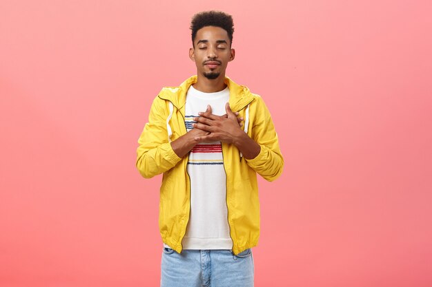 Attractive and stylish dark-skinned guy feeling warm and calm feelings inside holding hands on heart closing eyes and smiling with relieved comfort expression standing over pink wall in trendy outfit