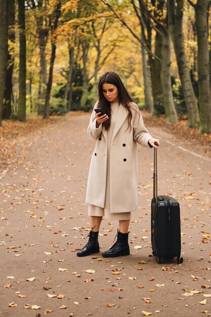 スーツケースと美しい秋の公園に立っている携帯電話を熱心に使用してコートを着た魅力的なスタイリッシュな女の子