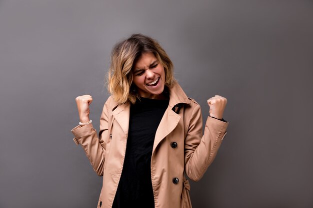 Attractive stylish girl with short stylish hairstyle in beige trench dancing with inspired face expression. Active young woman in casual autumn outfit having fun indoor and showing success