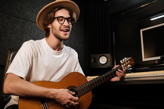 Привлекательный стильный мужчина поет и играет на гитаре в студии. Молодой музыкант записывает новую песню в современной звуковой студии