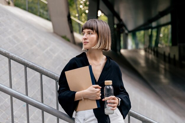 Attractive stylish shorthaired woman in blouse is standing on business district Happy girl with straight hairstyle holding notebooks and water outside