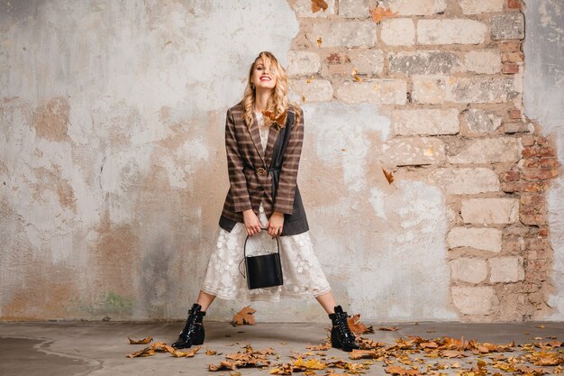 Attractive stylish smiling blonde woman in checkered jacket walking against wall in street