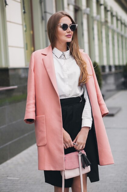 Attractive stylish smiling rich woman walking city street in pink coat spring fashion trend holding purse, elegant style, wearing sunglasses