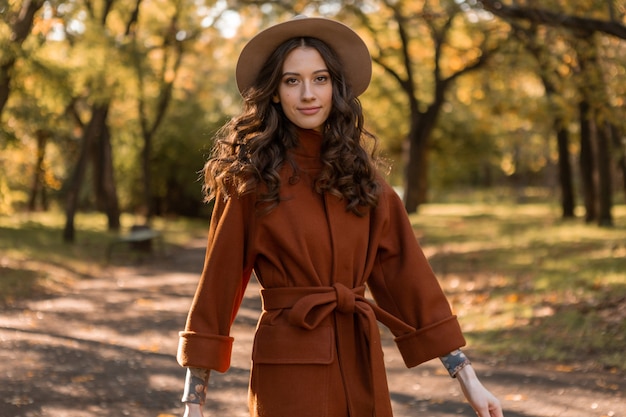 따뜻한 갈색 코트, 가을 유행 패션 스트리트 스타일을 입고 공원에서 산책하는 곱슬 머리를 가진 매력적인 세련된 웃는 마른 여자