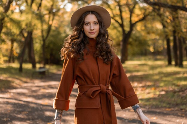Привлекательная стильная улыбающаяся тощая женщина с вьющимися волосами гуляет в парке, одетая в теплое коричневое пальто, осенний модный уличный стиль