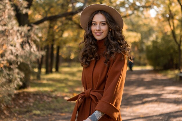 暖かい茶色のコート、秋の流行のファッションストリートスタイルに身を包んだ公園を歩く巻き毛の魅力的なスタイリッシュな笑顔の細い女性