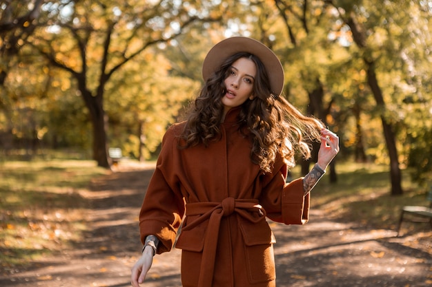 따뜻한 갈색 코트, 가을 유행 패션 스트리트 스타일을 입고 공원에서 산책하는 곱슬 머리를 가진 매력적인 세련된 웃는 마른 여자