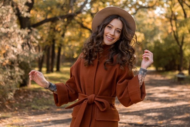 Привлекательная стильная улыбающаяся тощая женщина с вьющимися волосами гуляет в парке, одетая в теплое коричневое пальто, осенний модный уличный стиль