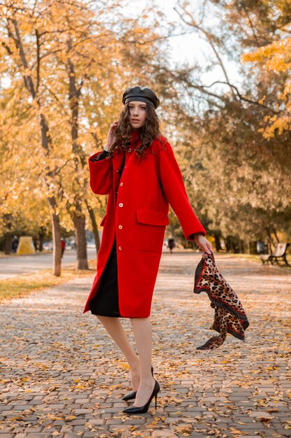 暖かい赤いコートの秋の流行のファッション、ストリートスタイル、ベレー帽の帽子とヒョウ柄のスカーフを身に着けて公園を歩く巻き毛の魅力的なスタイリッシュな笑顔の細い女性