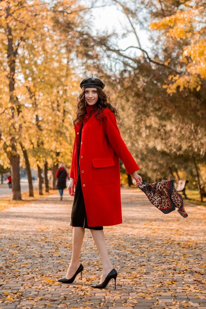 Привлекательная стильная улыбающаяся тощая женщина с вьющимися волосами гуляет в парке, одетая в теплое красное пальто осенней модной моды, уличного стиля, в шляпе-берете и шарфе с леопардовым принтом