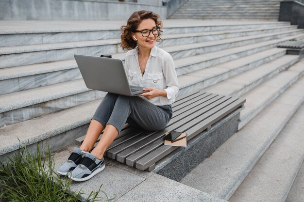 Привлекательная стильная улыбающаяся женщина в очках, работающих с набором текста на ноутбуке