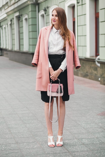 Attractive stylish smiling woman walking city street in pink coat