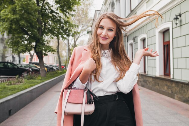 Attractive stylish smiling woman walking city street in pink coat