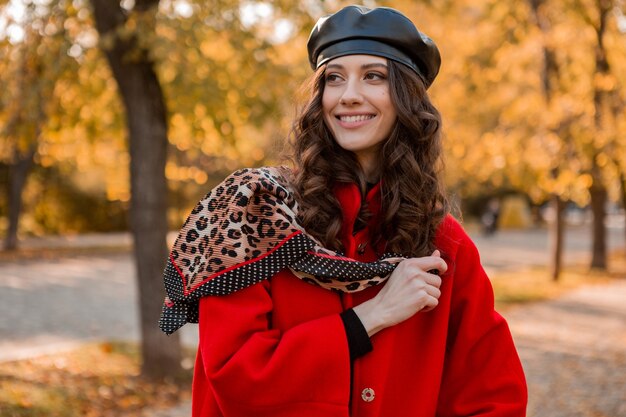 Attractive stylish smiling woman with curly hair walking in park dressed in warm red coat autumn trendy fashion, street style, wearing beret hat and leopard printed scarf