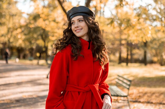 Attractive stylish smiling woman with curly hair walking in park dressed in warm red coat autumn trendy fashion, street style, wearing beret hat