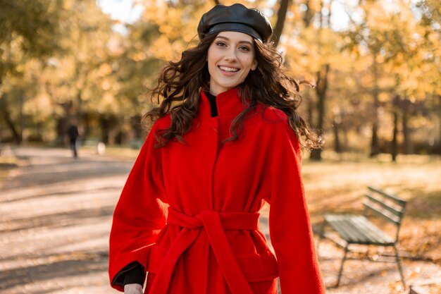 Привлекательная стильная улыбающаяся женщина с вьющимися волосами гуляет в парке, одетая в теплое красное пальто осенней модной моды, уличного стиля, в шляпе-берете