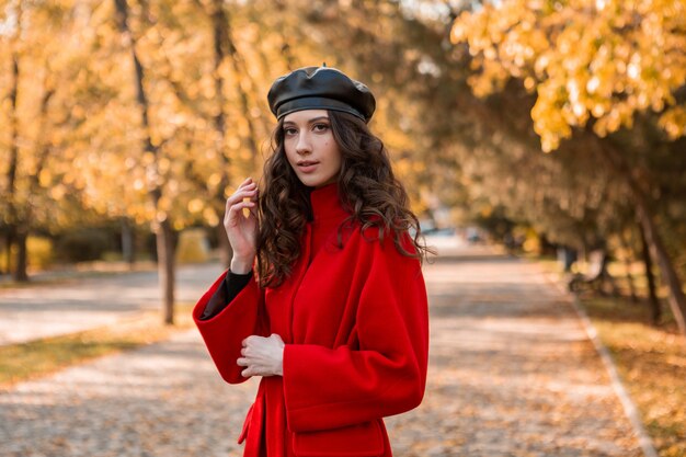 Attractive stylish smiling woman with curly hair walking in park dressed in warm red coat autumn trendy fashion, street style, wearing beret hat