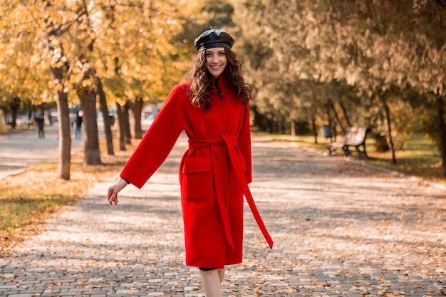 Attractive stylish smiling woman with curly hair walking in park dressed in warm red coat autumn trendy fashion, street style, wearing beret hat