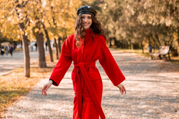 暖かい赤いコート秋の流行のファッション、ストリートスタイル、ベレー帽の帽子をかぶって公園を歩く巻き毛の魅力的なスタイリッシュな笑顔の女性