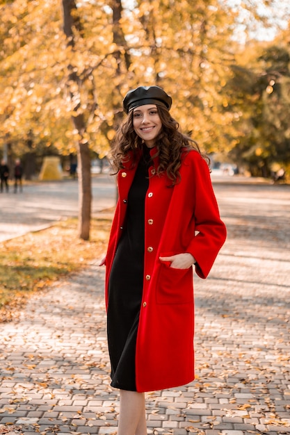 Attractive stylish smiling woman with curly hair walking in park dressed in warm red coat autumn trendy fashion, street style, wearing beret hat