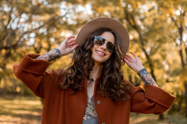 Привлекательная стильная улыбающаяся женщина с вьющимися волосами гуляет в уличном парке, одетая в теплое коричневое пальто осенней модной моды, уличного стиля в шляпе и солнцезащитных очках