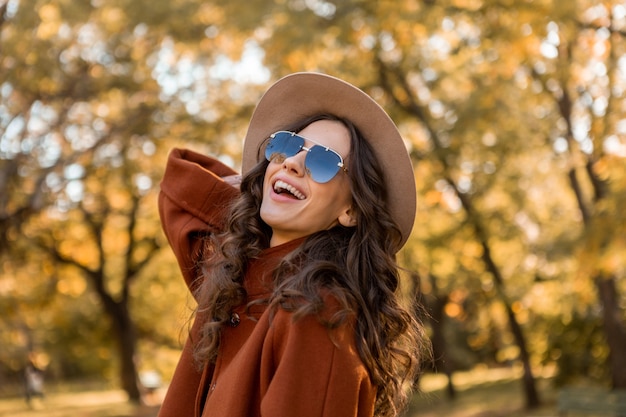 Attractive stylish smiling woman with curly hair walking in street park dressed in warm brown coat autumn trendy fashion, street style wearing hat and sunglasses