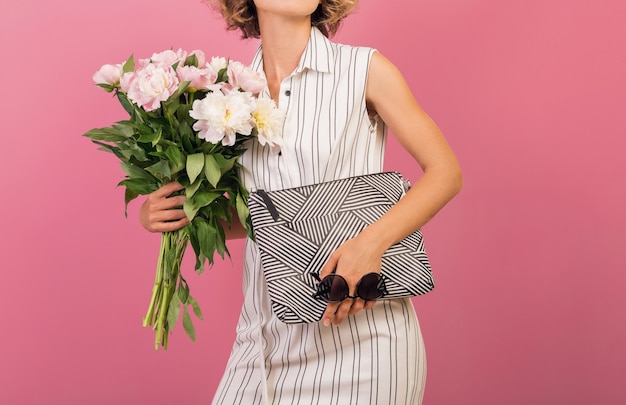 Attractive stylish woman in elegant white striped dress on pink studio background emotional face expression, surprised, handbag, flower bouquet, funny, curly hairstyle, fashion summer trend accessory