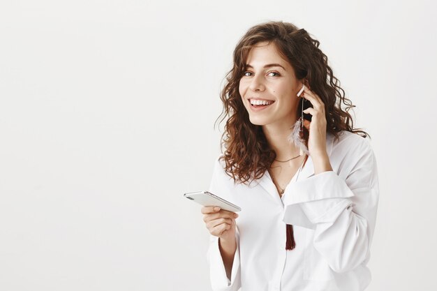 Attractive stylish woman listening music in wireless earphones, holding smartphone