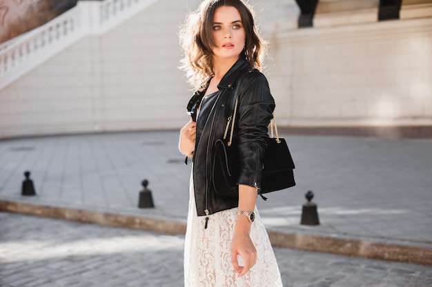 Attractive stylish woman walking in street in fashionable outfit, holding suede handbag, wearing black leather jacket and white lace dress, spring autumn style, turning around in motion