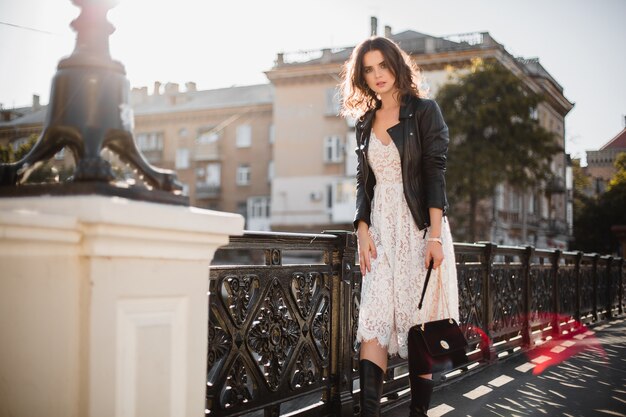 Attractive stylish woman walking in street in fashionable outfit holding suede handbag wearing black leather jacket and white lace dress, spring autumn style