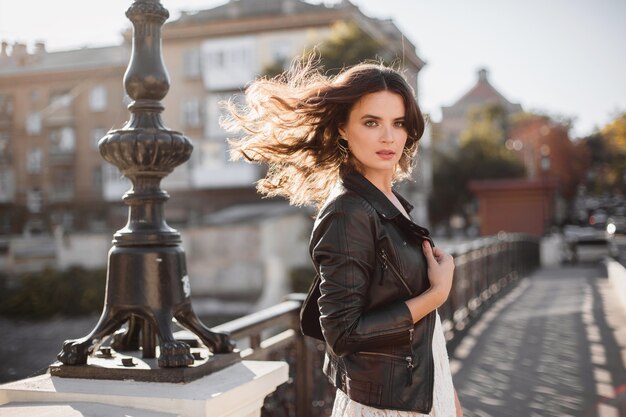 Attractive stylish woman walking in street in fashionable outfit waving hair in wind wearing black leather jacket and white lace dress spring autumn style