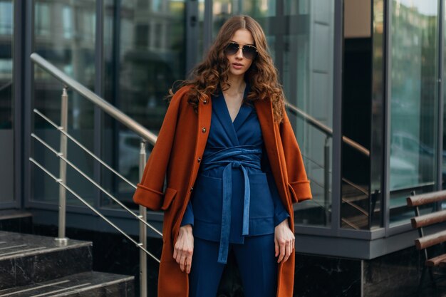 Attractive stylish woman with walking in urban city business street dressed in warm brown coat and blue suit, spring autumn trendy fashion street style, wearing sunglasses