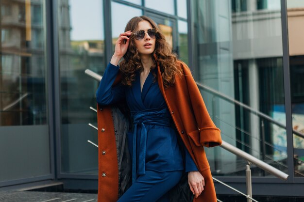 Attractive stylish woman with walking in urban city business street dressed in warm brown coat and blue suit, spring autumn trendy fashion street style, wearing sunglasses