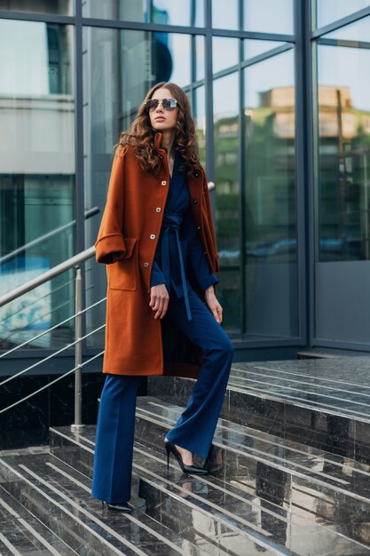 Attractive stylish woman with walking in urban city business street dressed in warm brown coat and blue suit, spring autumn trendy fashion street style, wearing sunglasses