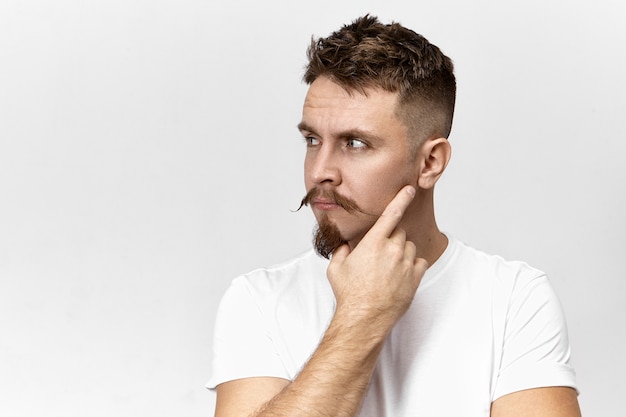 Attractive stylish young bearded guy with mustache thinking over something, holding hand on his face, posing against blank studio wall background with copy space for your advertising information