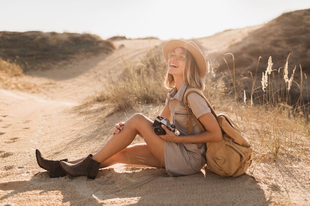 Attractive stylish young woman in khaki dress in desert, traveling in Africa on safari, wearing hat and backpack, taking photo on vintage camera