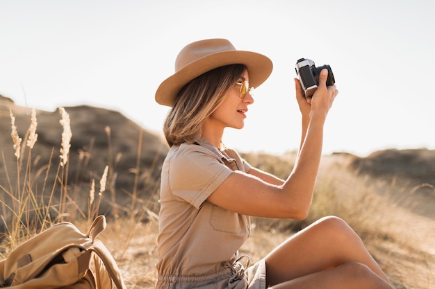 Attractive stylish young woman in khaki dress in desert, traveling in Africa on safari, wearing hat and backpack, taking photo on vintage camera