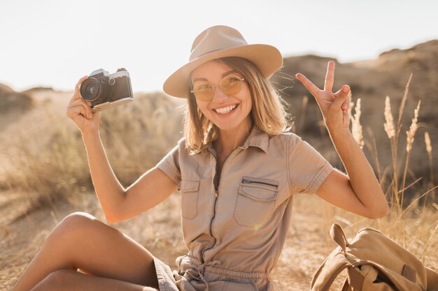 Attractive stylish young woman in khaki dress in desert, traveling in Africa on safari, wearing hat and backpack, taking photo on vintage camera