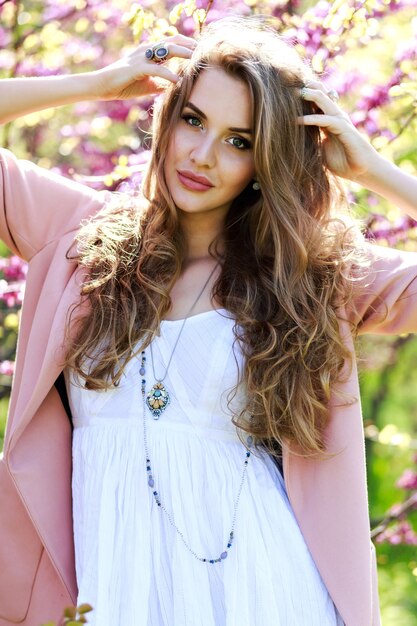 Привлекательная стильная молодая женщина в легком белом платье, розовом пальто, с длинными волосами, гуляет в саду с цветущей сакурой