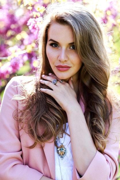 Attractive stylish young woman in light white dress, pink coat, with long hair walking in garden with blooming sakura