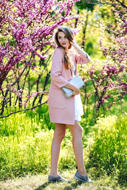 Привлекательная стильная молодая женщина в легком белом платье, розовом пальто, с длинными волосами, гуляет в саду с цветущей сакурой